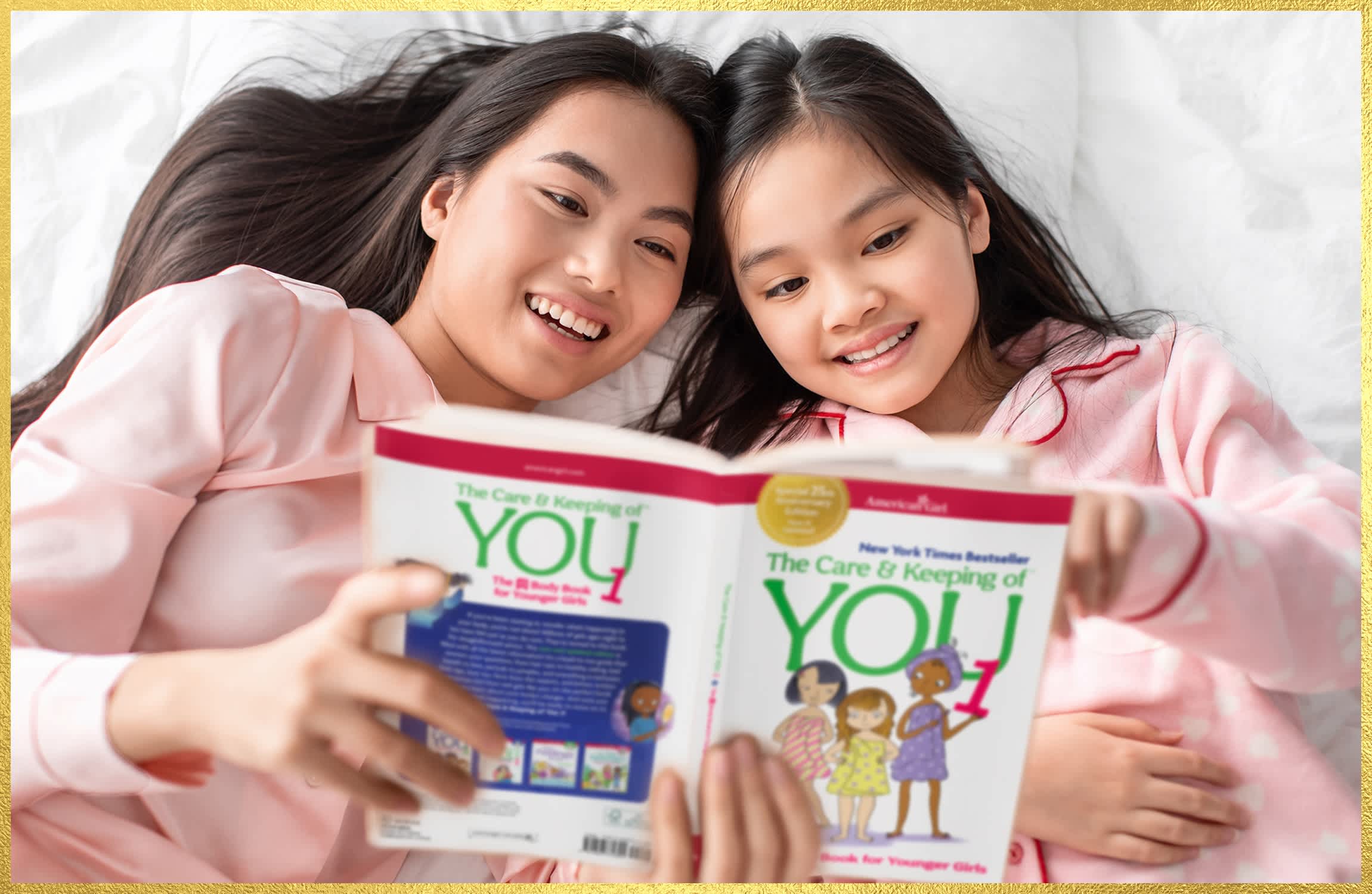 A younger female presenting person and a seemingly older female presenting person wearing matching pink pajamas lying on a bed while reading The Care & Keeping of You book from American Girl.