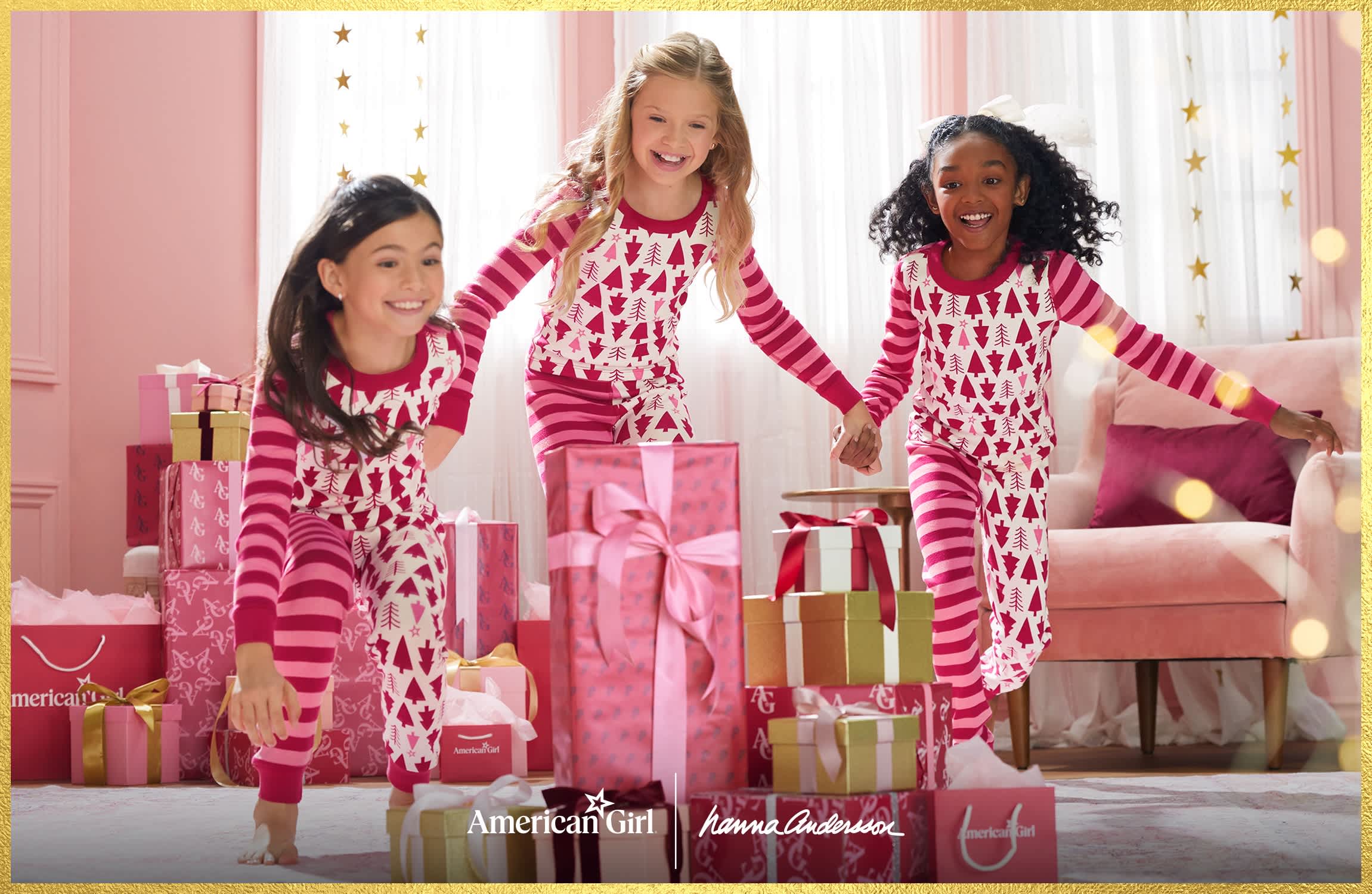 Three children wearing matching red, pink, and white American Girl x Hanna Andersson holiday pajamas run and play in a room decorated with wrapped gifts and a pink sofa.