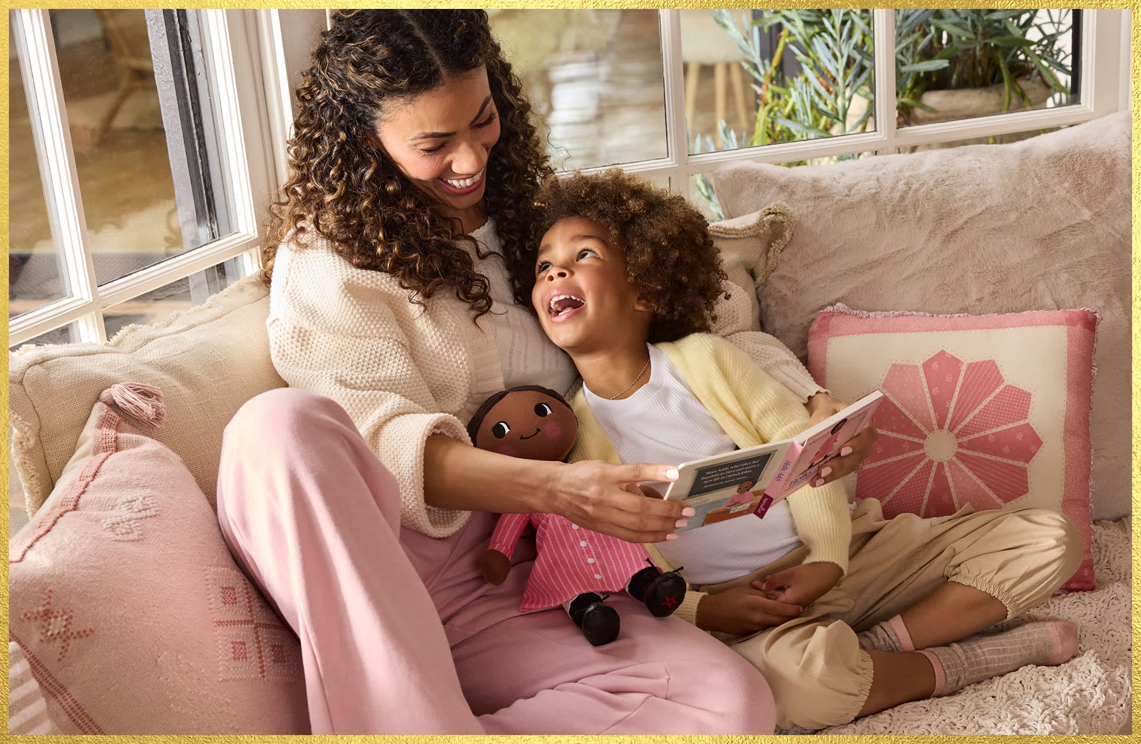 An adult and child sit on a cozy sofa reading a storybook together, while the child holds an American Girl Addy plush doll dressed in a pink outfit.