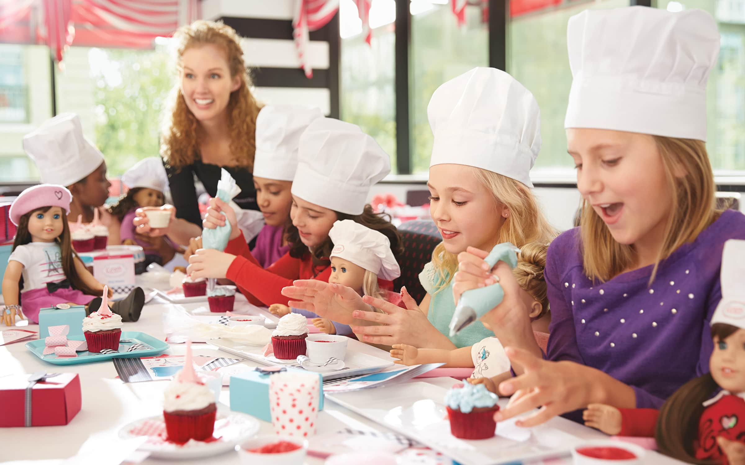 Young girls with chef hats decorating their cupcakes next to their dolls